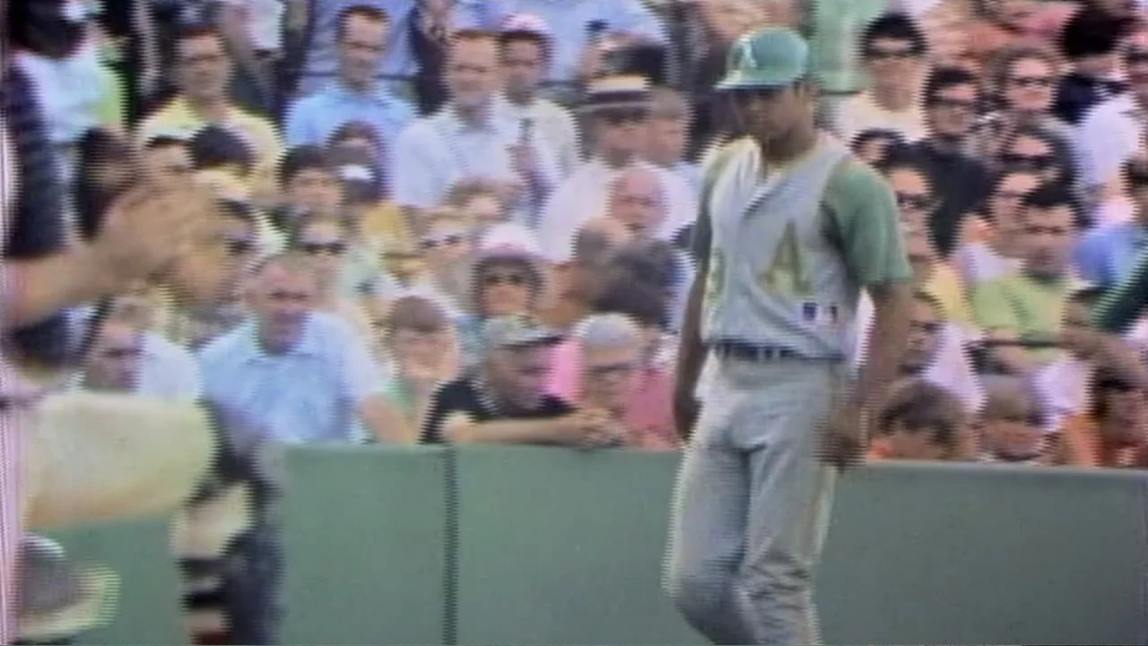 Reggie (2023) movie still: medium — A baseball player walks across a green field with spectators in the background. The scene features vintage…