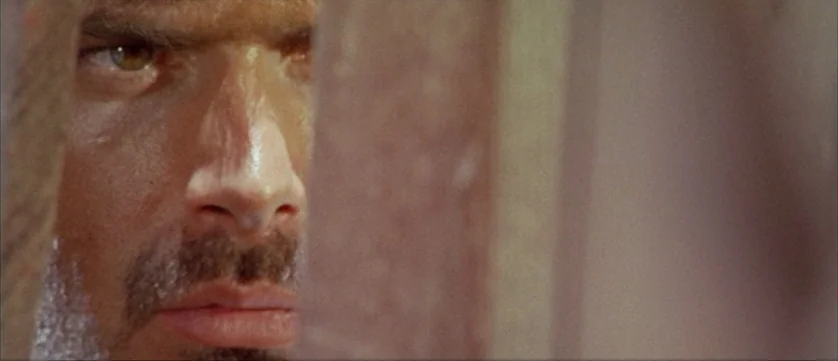 Django Kill (1967) movie still: closeup — A man's face is partially obscured by a textured, vertical brown barrier. He looks through the narrow…