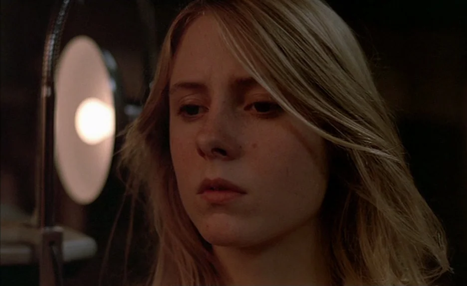 Black Moon (1975) movie still: closeup — A dimly lit space features a glowing vanity mirror lamp in the foreground. A young woman with blonde hair…