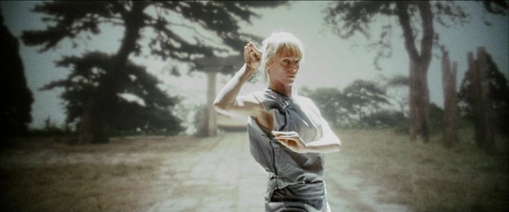 Kill Bill Vol. 2 (2004) movie still: medium — An outdoor courtyard with large trees and a stone structure in the distance. A woman in a grey tunic…