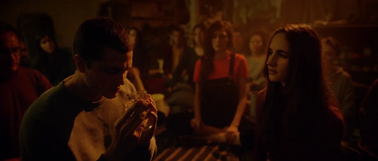 Love (2015) movie still: medium — A group of young adults sit on the floor in a dimly lit, cluttered basement. A man holds a glass in front of his…
