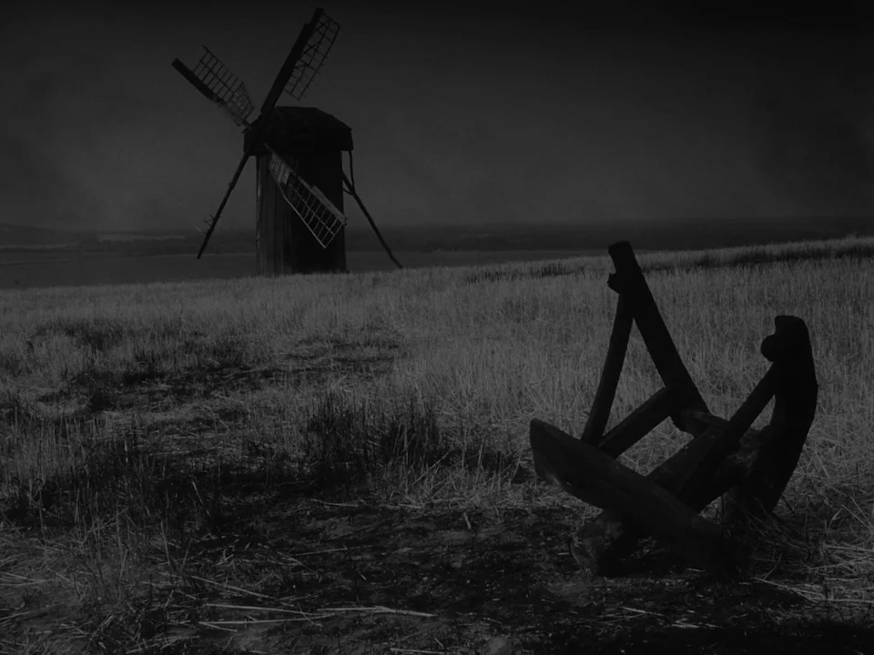 Ivan’s Childhood (1962) movie still: wide — A dark, grassy field extends toward a distant, weathered windmill under a clouded sky. A heavy wooden…