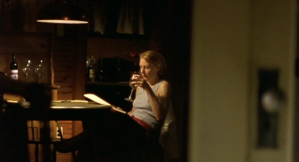 The Station Agent (2003) movie still: medium — A dimly lit room with dark wood walls features a circular dining table. A woman sits at the table,…