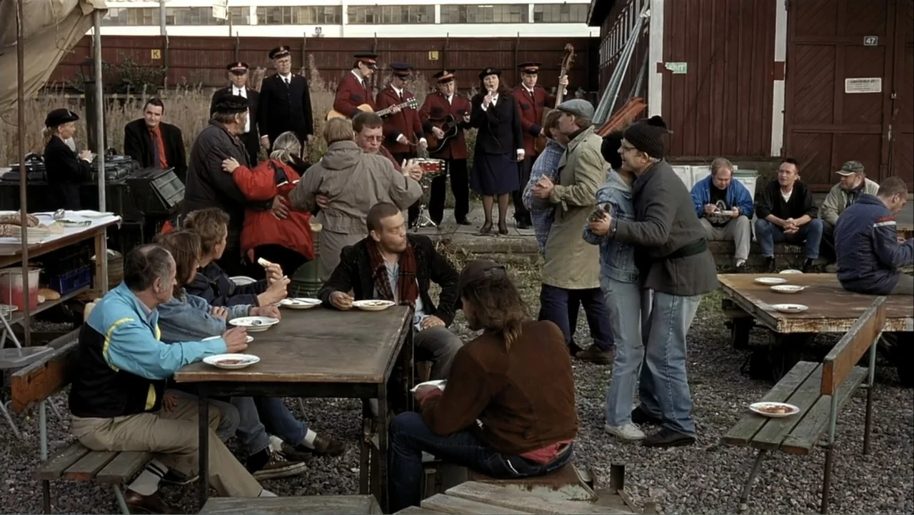 The Man Without a Past (2002) movie still: wide — A gravel industrial site features a wooden building and metal fence background. Multiple people…
