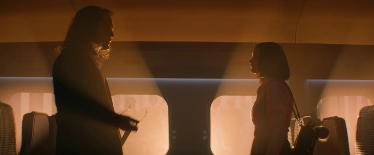 Bullet Train (2022) movie still: medium — The interior of a brightly lit train carriage features orange-hued windows along the walls. Two people…