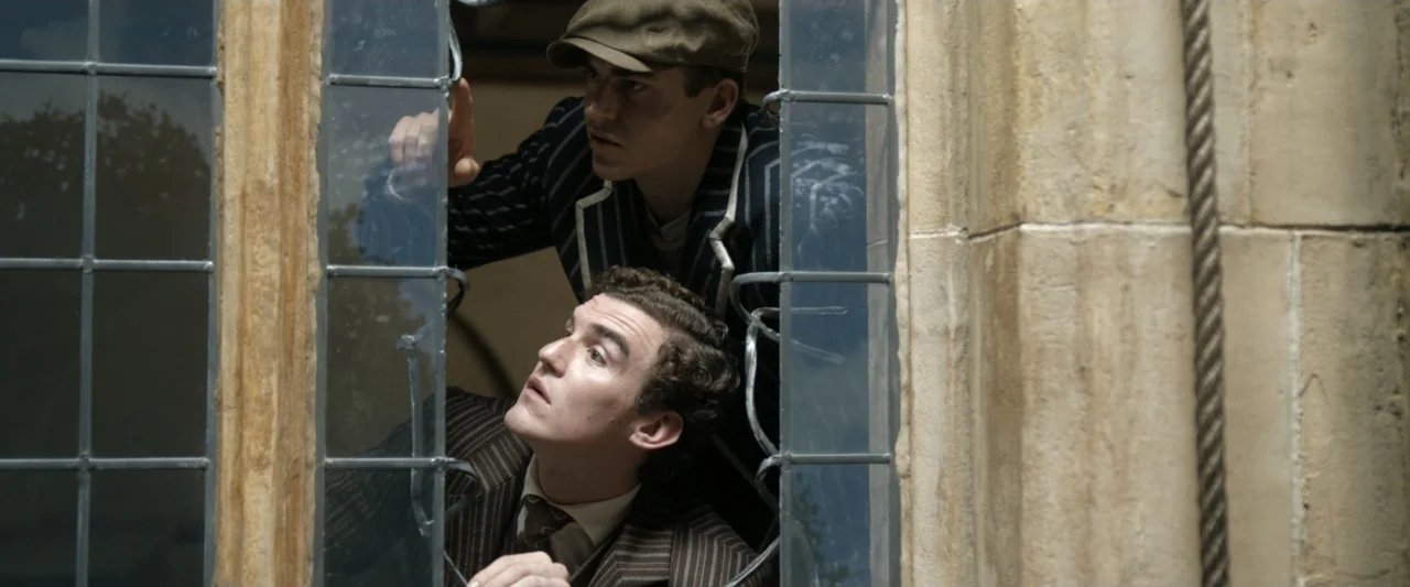 Young Sherlock Holmes (1985) movie still: medium — A stone building exterior with leaded glass windows and weathered masonry textures. Two men in…