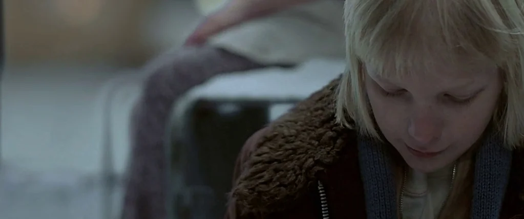 Let The Right One In (2008) movie still: closeup — A dimly lit indoor space with a neutral-colored background. A young blonde girl looks downward…