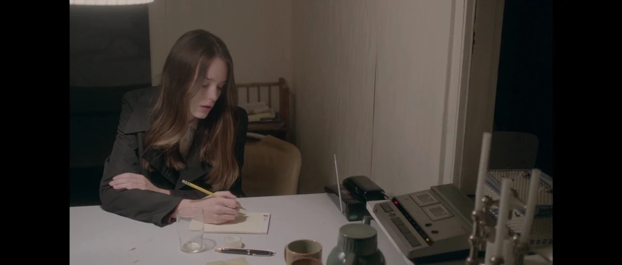 Nymphomaniac Vol 1 (2013) movie still: medium — A woman sits at a white laminate table in a dimly lit room. She uses a yellow pencil to write on a…