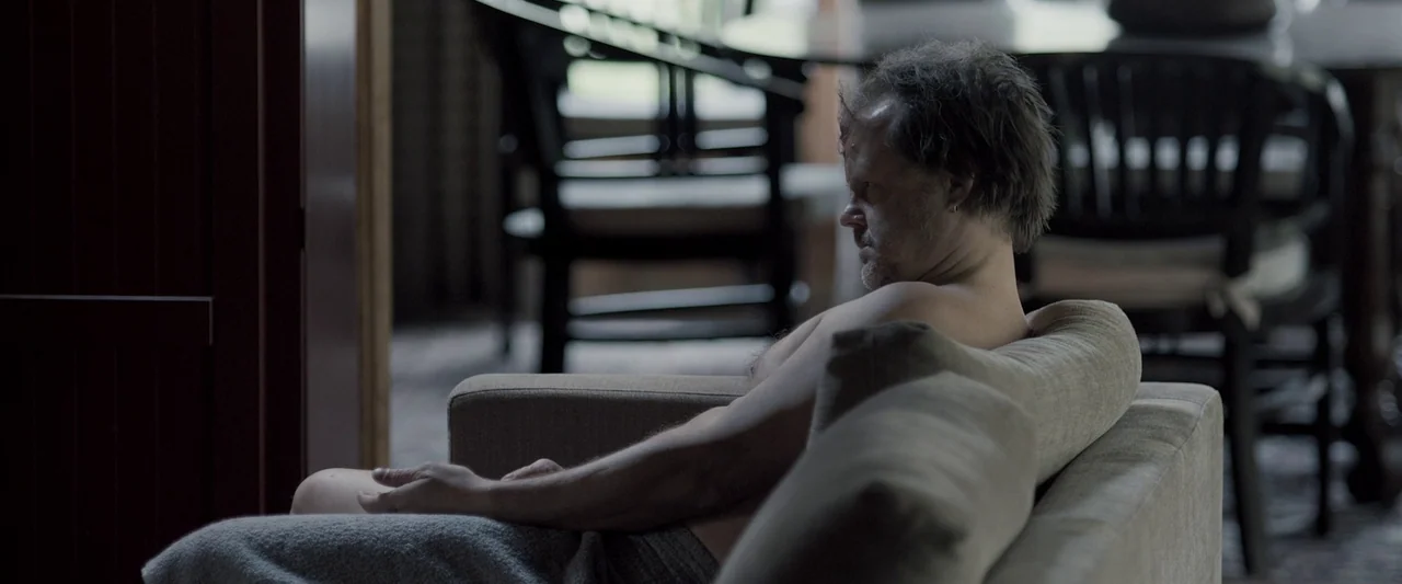 You’re Next (2013) movie still: medium — A man sits on a beige sofa in a dimly lit room. He is shirtless and partially covered by a grey blanket.