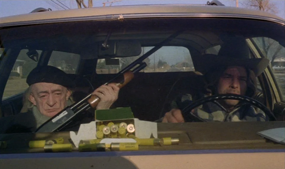 Stroszek (1977) movie still: medium — Two men sit inside a vehicle during the day with shotgun shells visible.