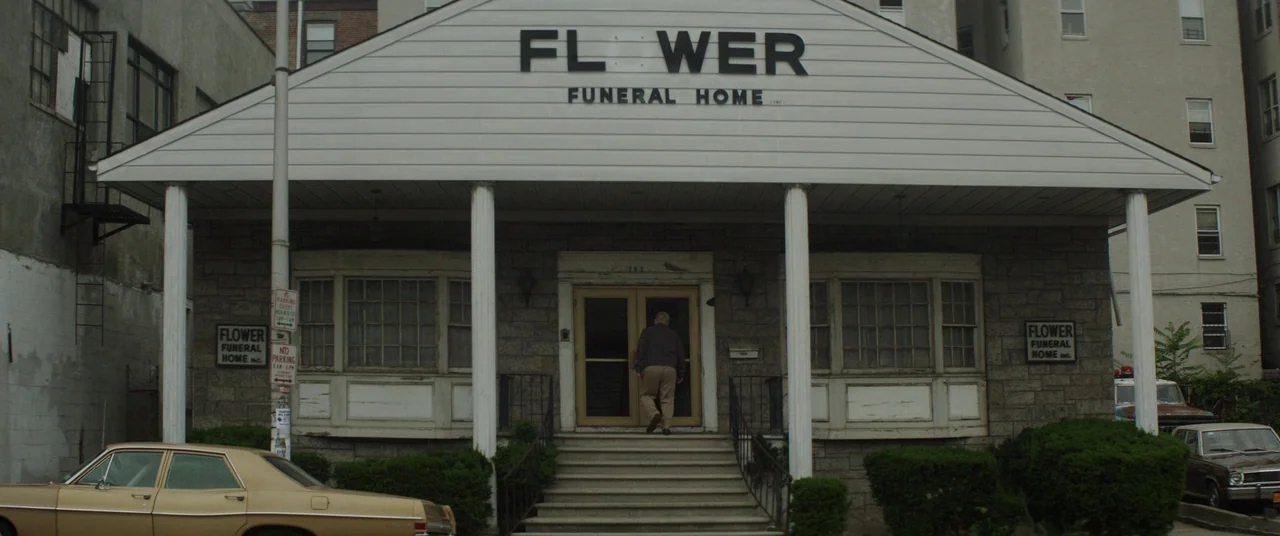God’s Pocket (2014) movie still: wide — A low-rise building with white siding and stone accents sits at street level. A man walks up the stairs to…