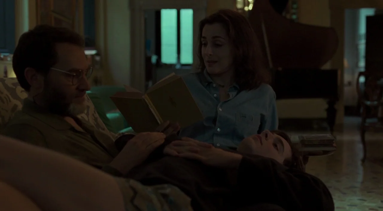 Call Me By Your Name (2017) movie still: medium — A dimly lit room with an upright piano in the background. A woman reads a book aloud to a man and…