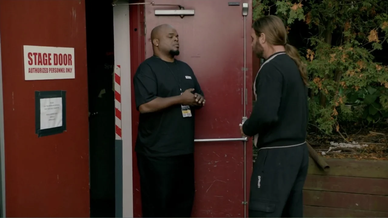 Clear History (2013) movie still: fullbody — A red metal stage door is situated in an outdoor area with trees nearby. A security guard speaks to a…