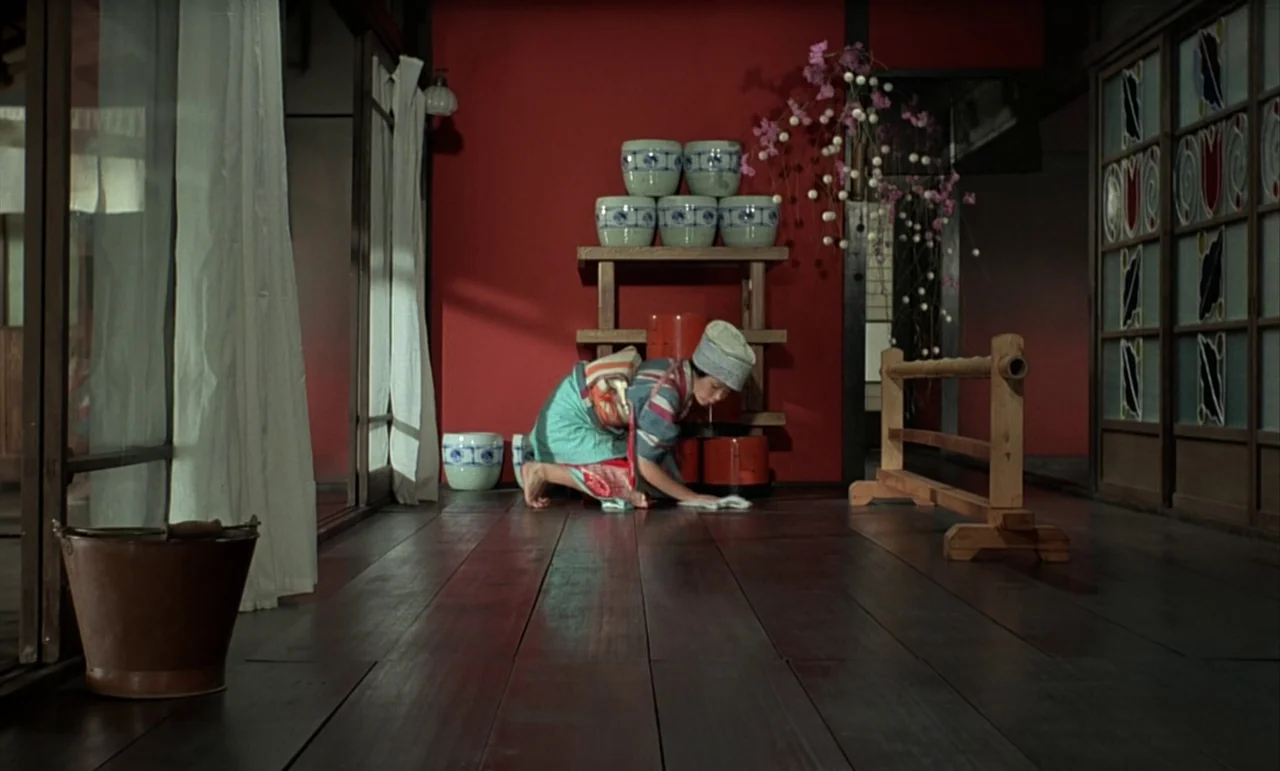 In The Realm of the Senses (1976) movie still: wide — A room in a traditional Japanese home features a vivid red wall and dark flooring. A woman in…
