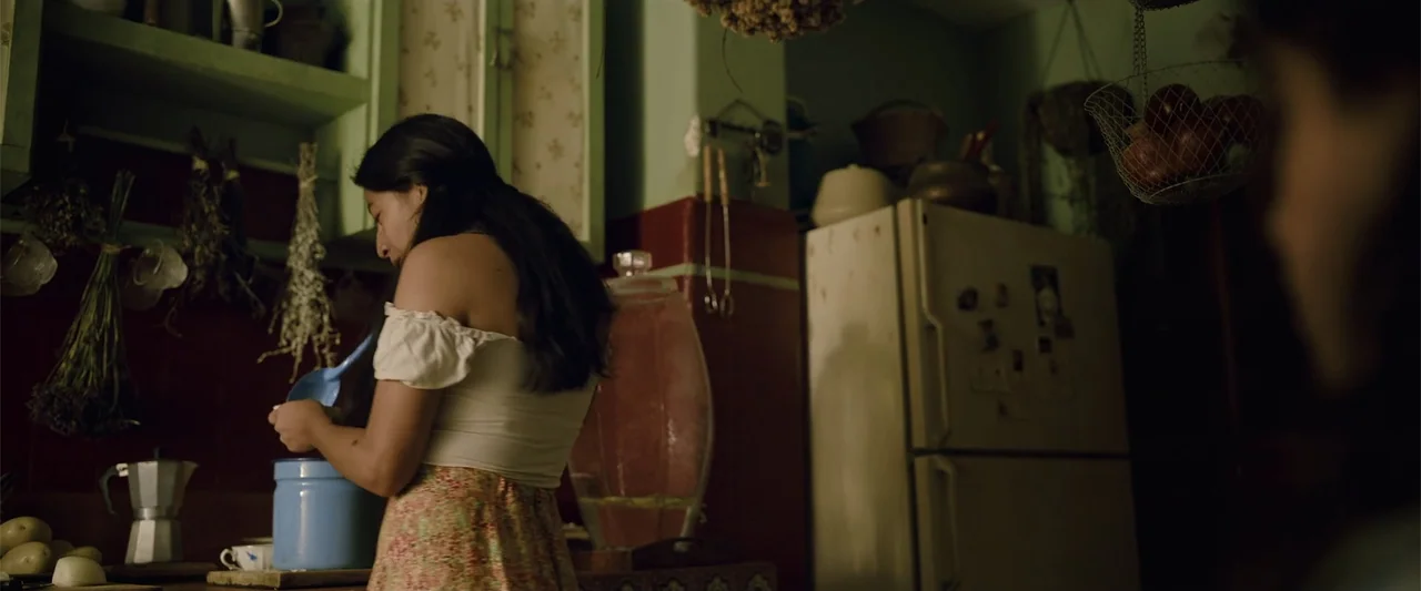 Evil Eye (2020) movie still: medium — A cluttered residential space features vintage cabinets and a refrigerator covered in magnets. A young woman…
