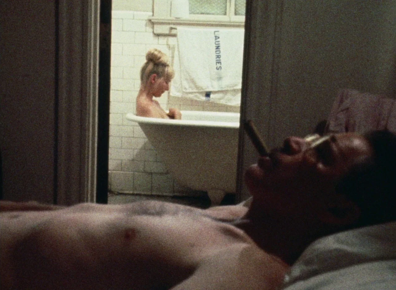 Wanda (1970) movie still: medium — A dark, cramped bedroom looks through a doorway into a lit tiled bathroom. A shirtless man smokes in bed while a…