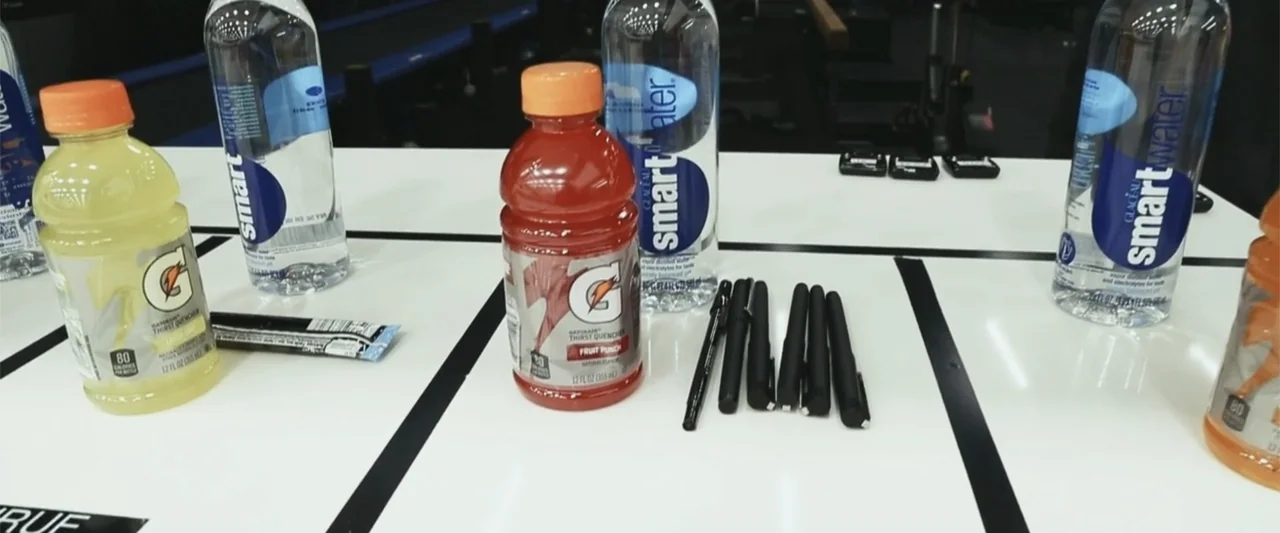 Giannis: The Marvelous Journey (2024) movie still: medium — A white table surface features several beverage bottles and a row of black pens. The…