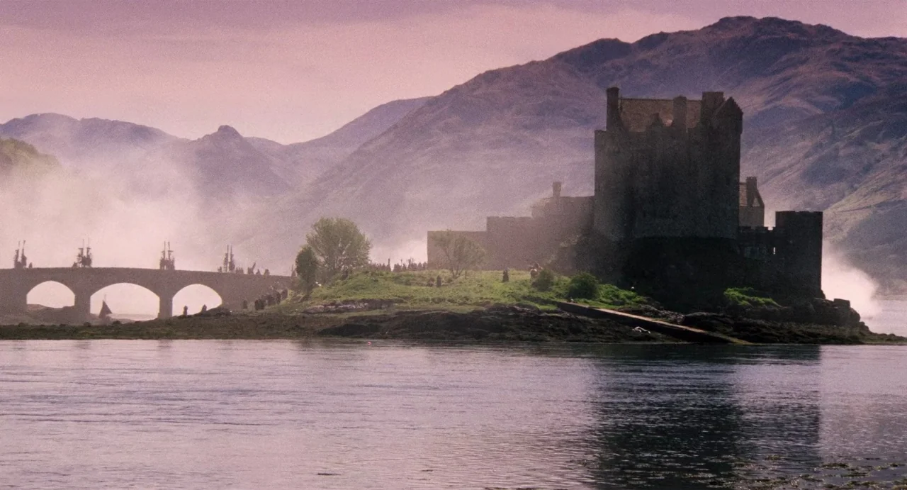 Highlander (1986) movie still: establishing — A stone castle sits on a small island surrounded by water with mountains behind. A bridge connects the…