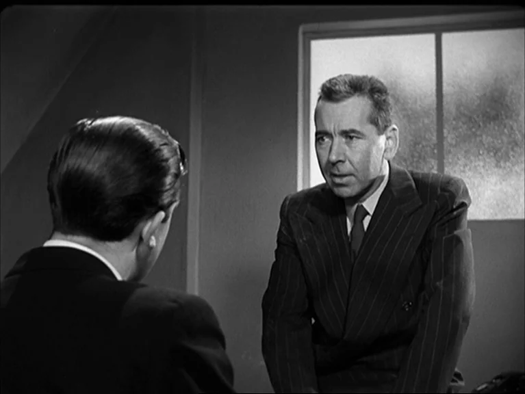 The Sleeping Tiger (1954) movie still: medium — A stark room features a frosted glass window and plain wall surfaces. A man in a dark pinstripe suit…