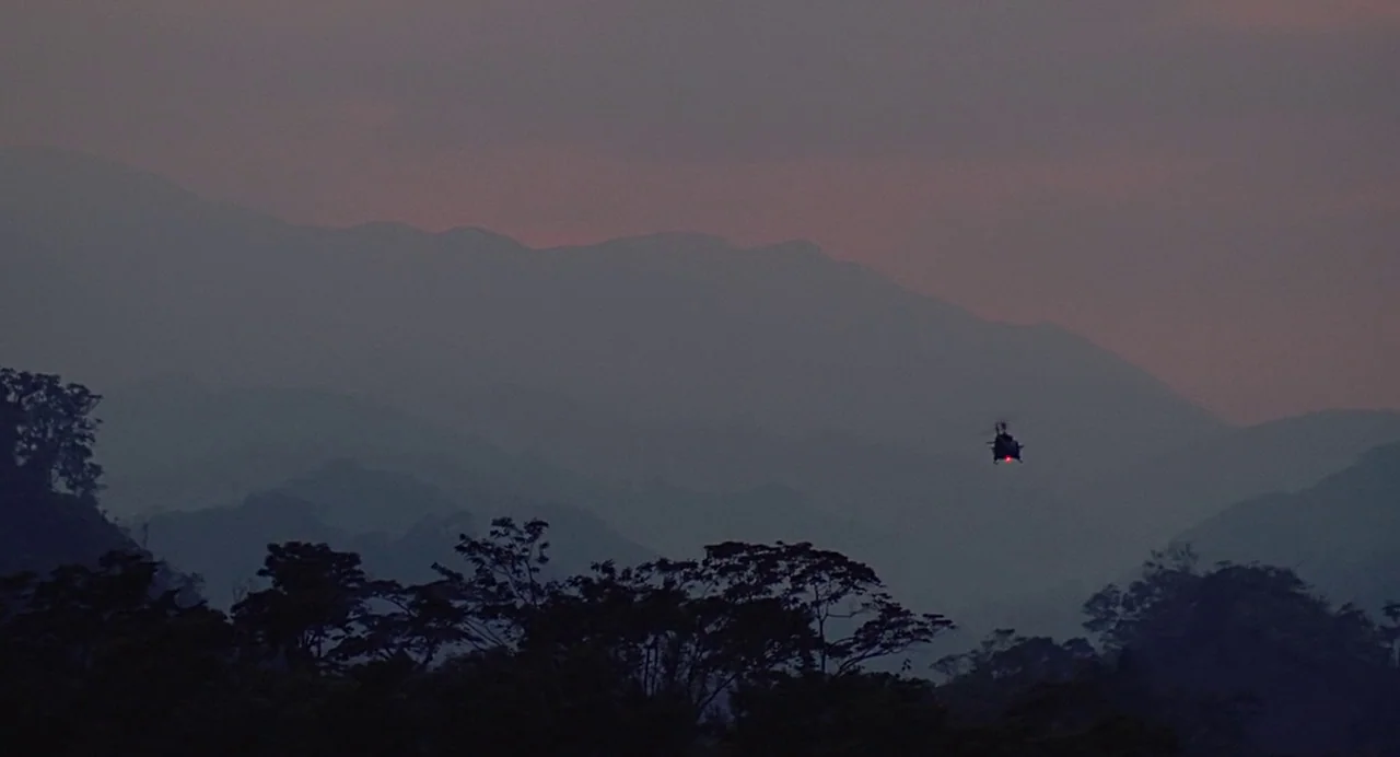 Predator (1987) movie still: establishing — A distant helicopter moves across the hazy silhouette of layered mountain ranges at twilight. Thick…