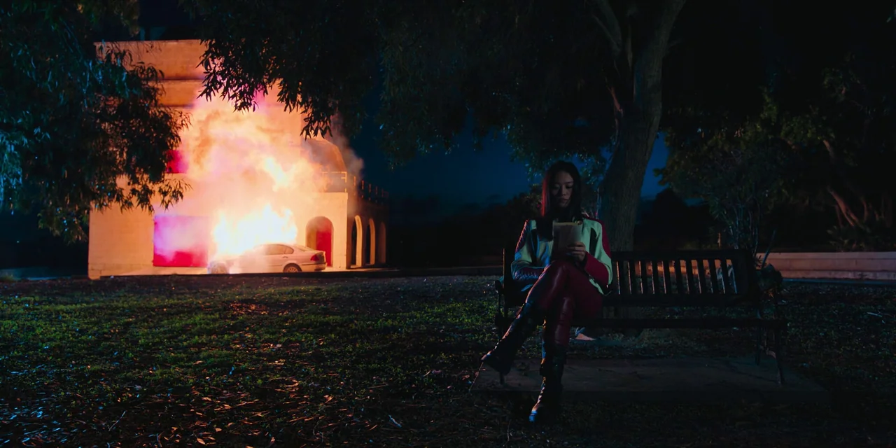 Sniper: G.R.I.T. (2023) movie still: wide — A park at night with a building and car engulfed in flames in background. A young woman sits on a bench…