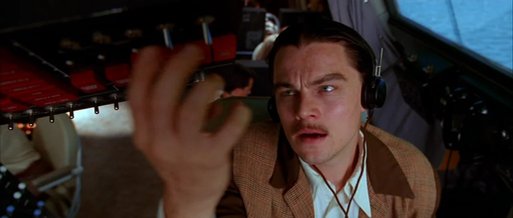 The Aviator (2004) movie still: medium — The interior of an airplane cockpit contains an array of red and black overhead switches. A man in a tweed…
