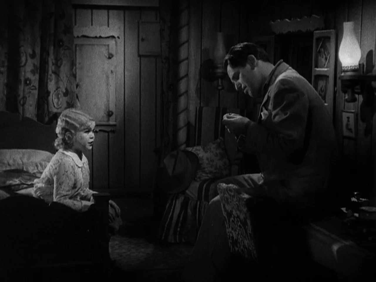 Freaks (1932) movie still: medium — A dimly lit room with wood-paneled walls features a bed and side furniture. An adult male sits while a young…