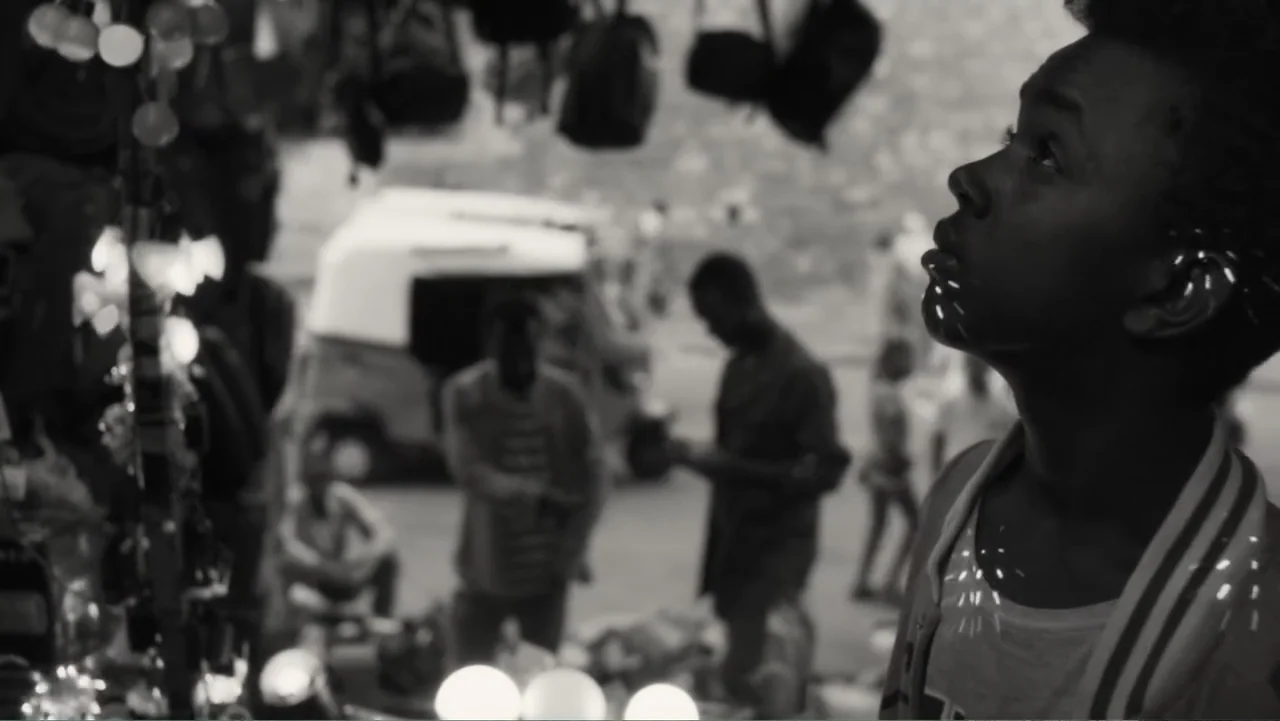 Faya Dayi (2021) movie still: closeup — A bustling night market scene with blurred figures and vehicles in the background. A young man stands in the…