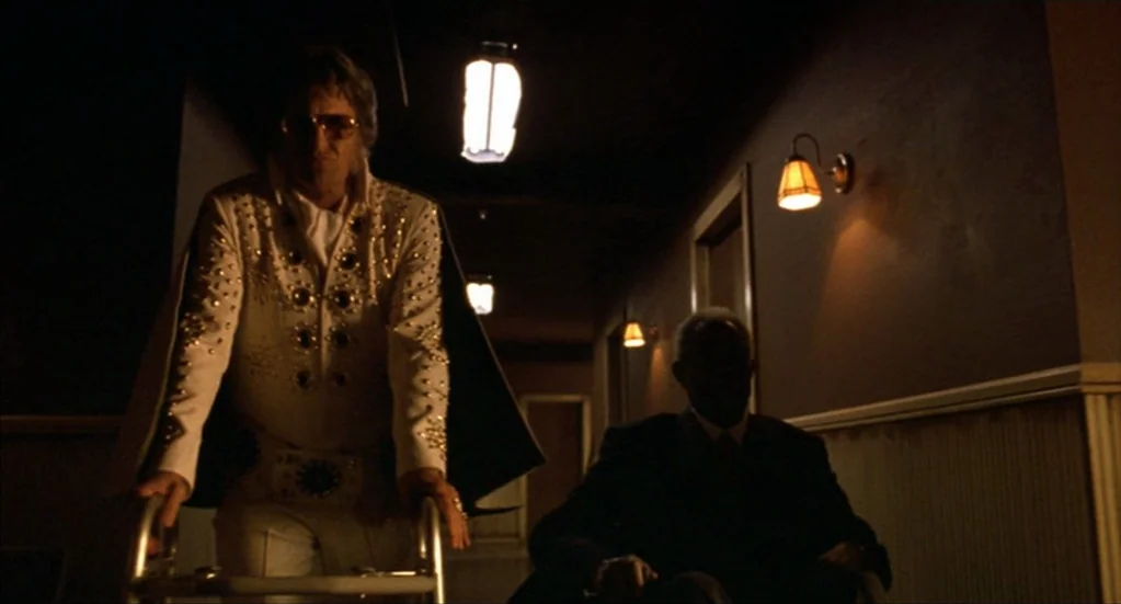 Bubba Ho-Tep (2002) movie still: medium — A dimly lit nursing home corridor features beige paneling and wall-mounted sconces. A person dressed as…