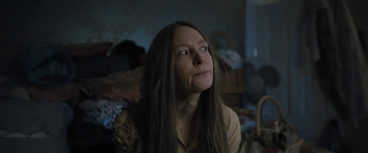 Longlegs (2024) movie still: closeup — A dimly lit room filled with miscellaneous boxes and household clutter. A middle-aged woman sits facing away…