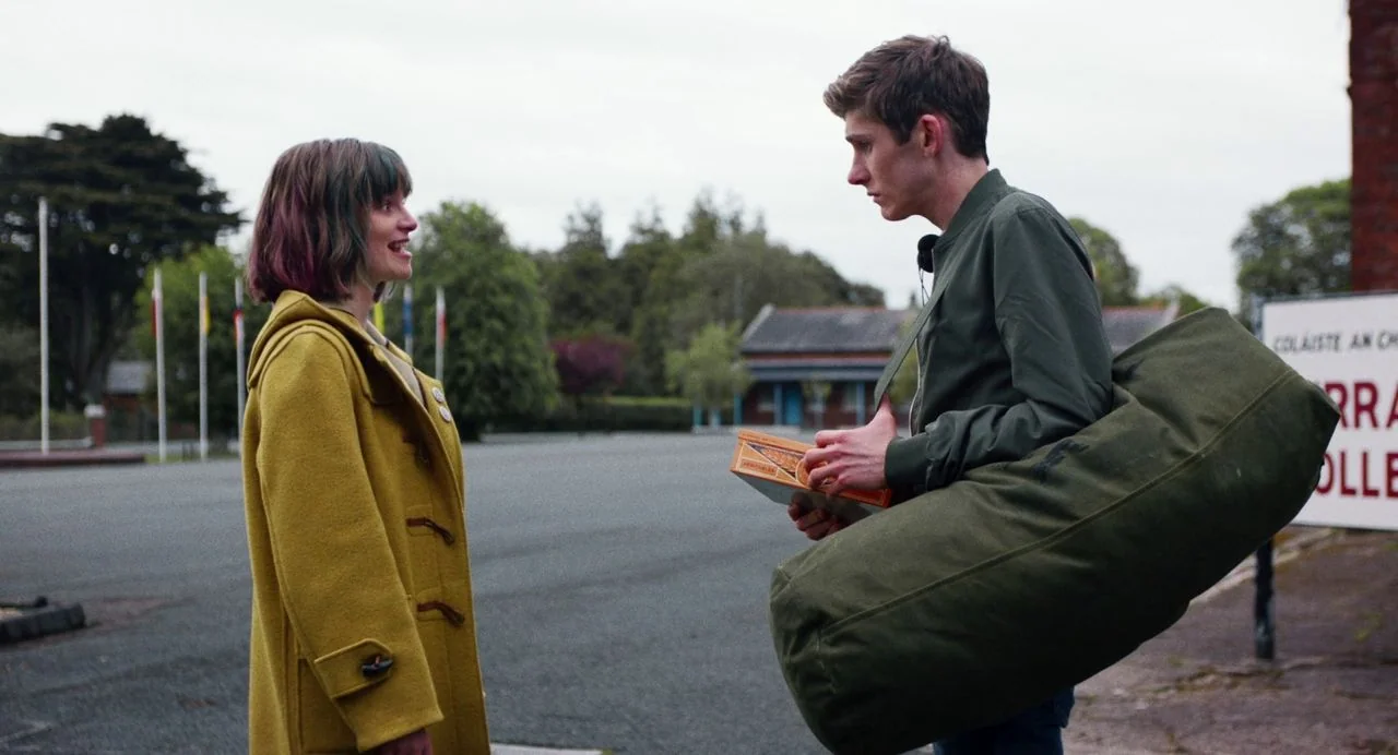 Dating Amber movie still: medium — A paved school entrance area features a line of flagpoles and a stone building. A teenage boy in a green jacket…