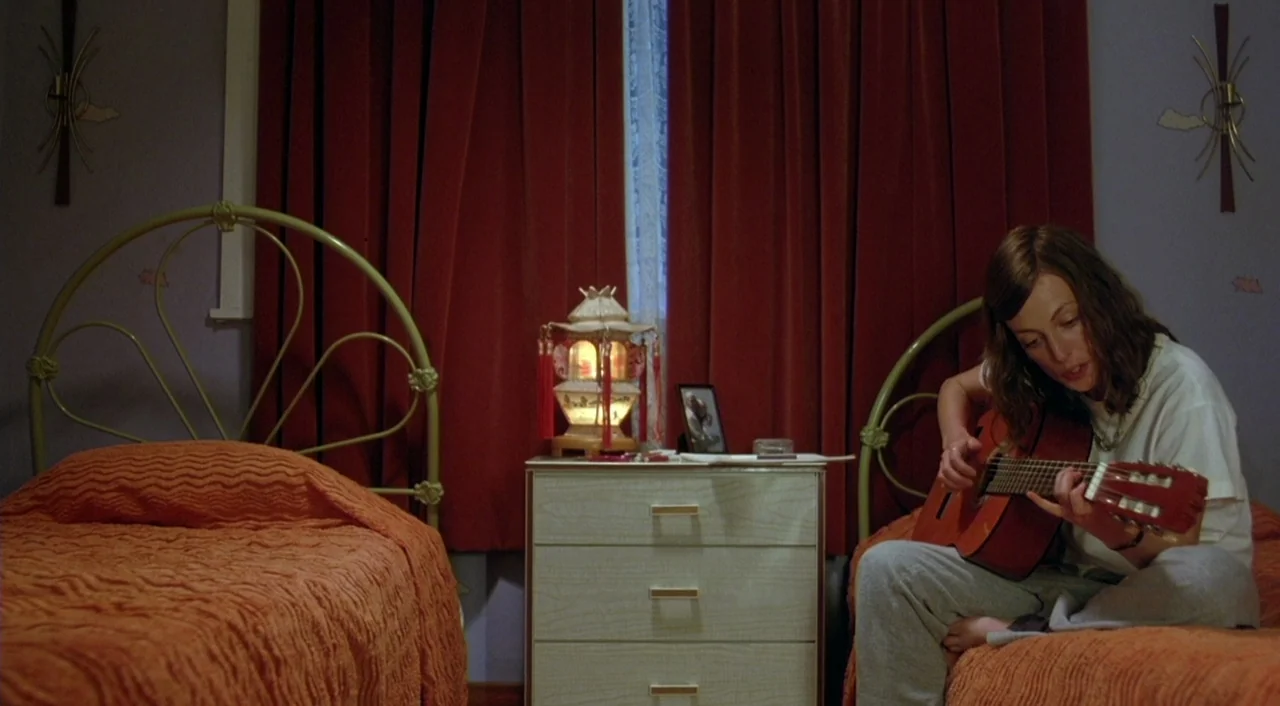 Eagle Vs. Shark movie still: medium — A room contains two twin beds with orange bedspreads and a central bedside table. A young woman sits on the…