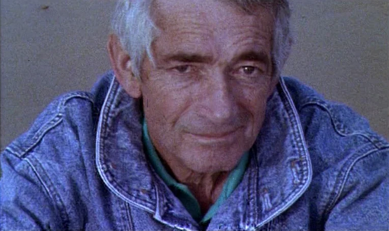 Jacquot de Nantes (1991) movie still: closeup — An older man sits on a sand-covered surface in outdoor natural light. He stares forward while…