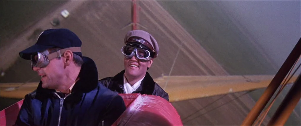 It Happened At The World’s Fair (1963) movie still: medium — Two men sit inside the red cockpit of a vintage biplane in flight. The landscape below…