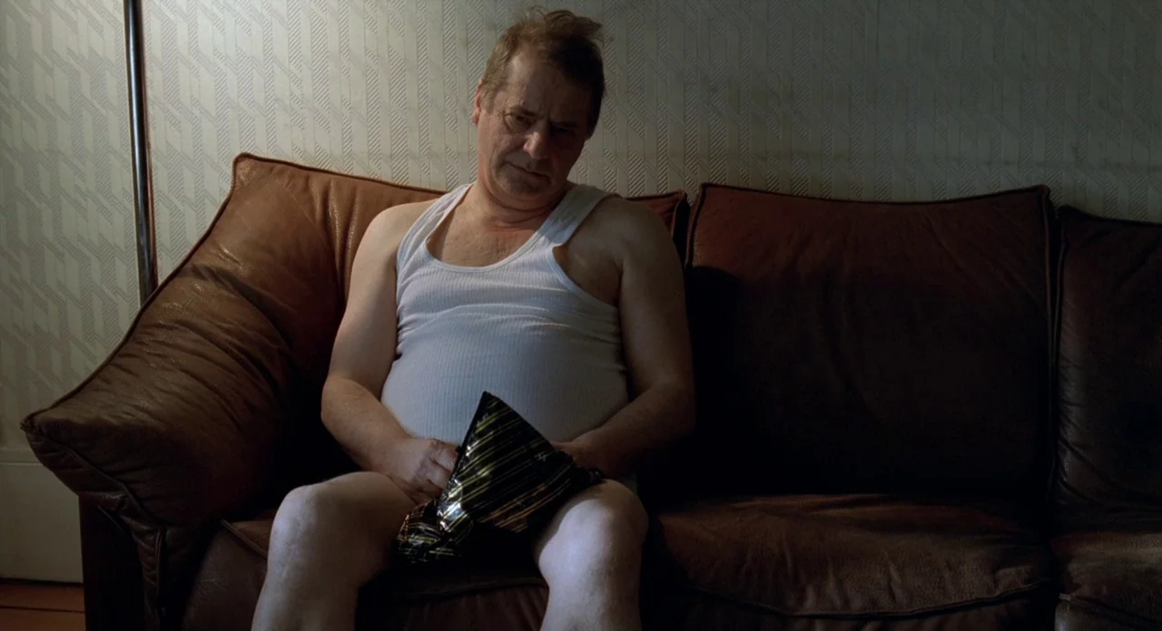 Read My Lips (2001) movie still: medium — A man sits on a brown leather couch in a room with patterned wallpaper. He holds a dark metallic snack bag…