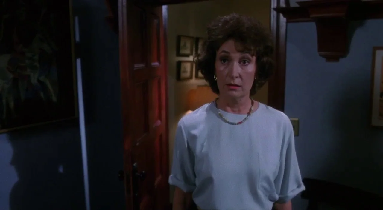 Heathers movie still: medium — A dark hallway with blue-grey walls features an open wooden door. A woman in a grey blouse stands in the entryway…