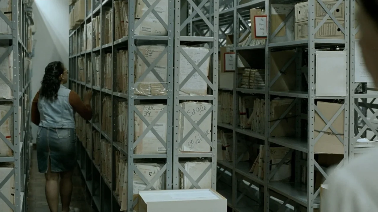 I'm Still Here (2024) movie still: medium — A dense archive room contains rows of tall metal shelving units filled with documents. A woman examines…