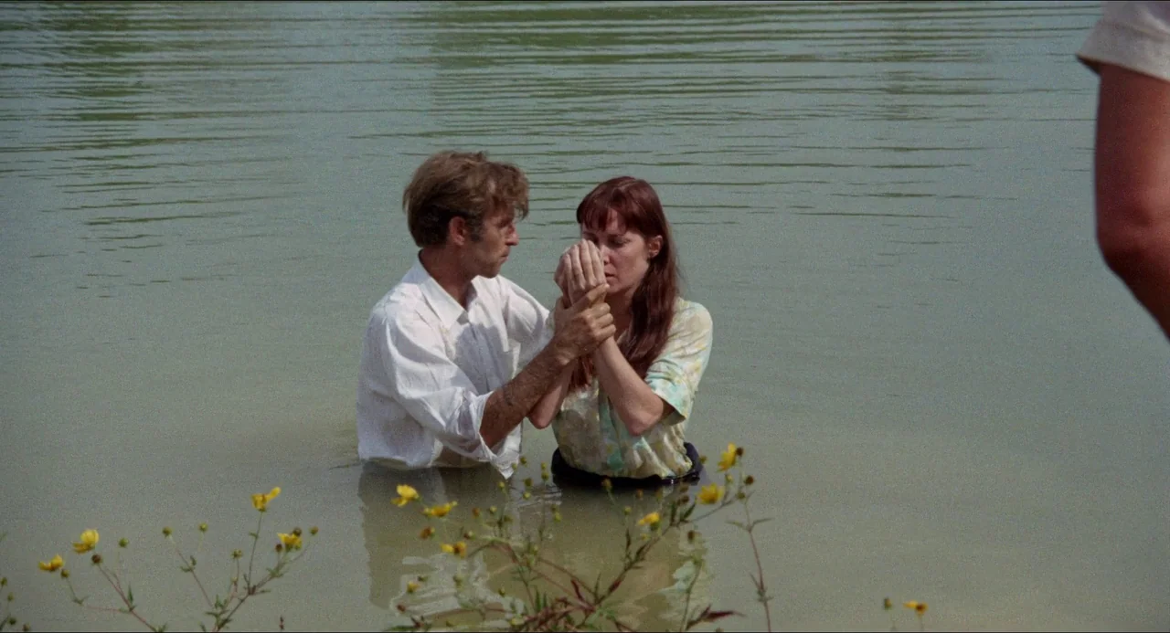 Medium Cool (1969) movie still: medium — A man and woman stand in murky, calm river water during the day. The man grasps the woman's joined hands in…
