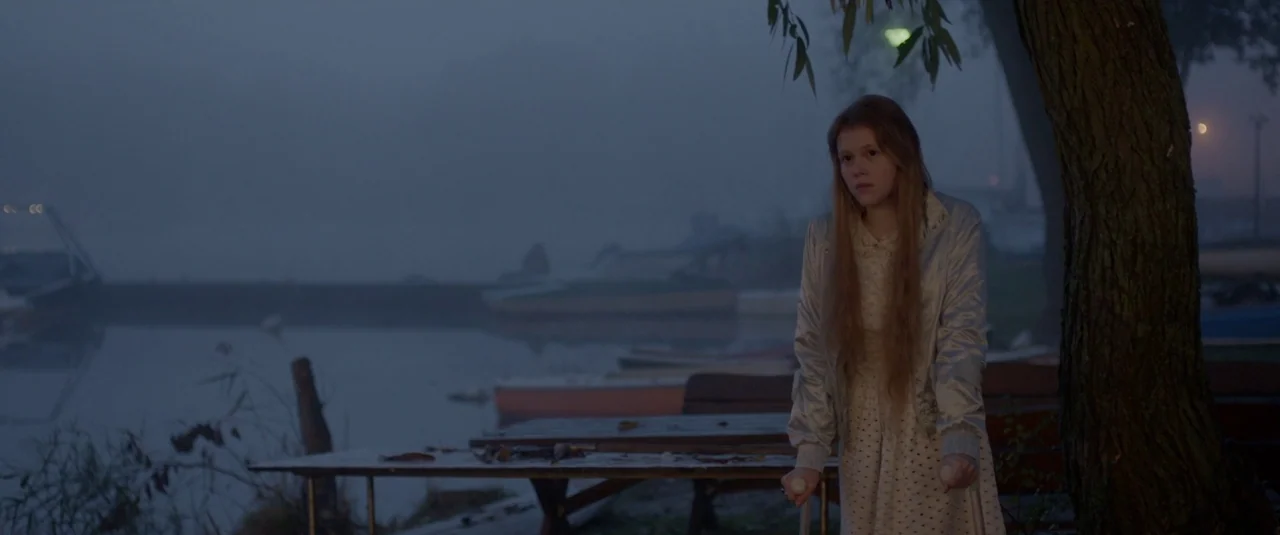 The Lure (2015) movie still: medium — A misty, dim waterfront at dusk features a large tree and docked boats nearby. A young girl with long hair…