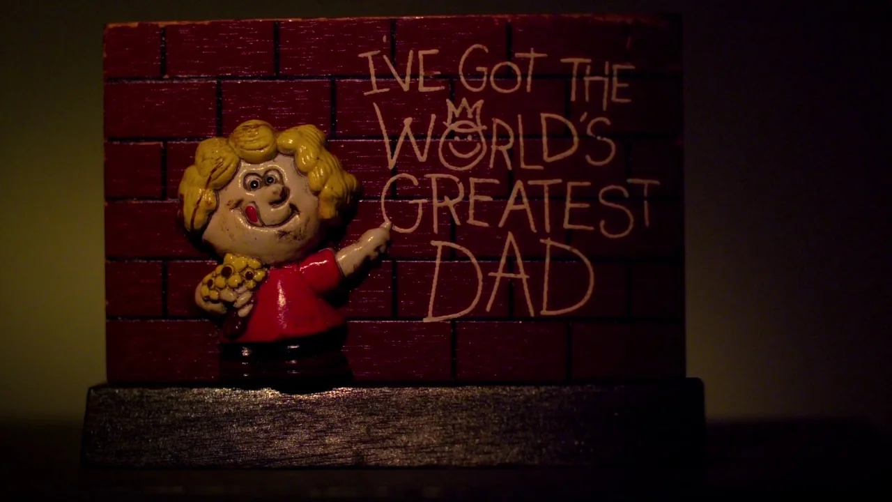 Suburban Gothic (2014) movie still: closeup — A small plastic figurine of a child stands before a faux-brick backdrop. The surface is inscribed with…