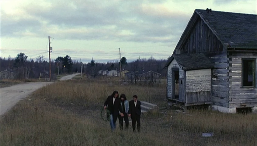 Dance Me Outside (1994) movie still: wide — A rural landscape features a derelict wooden cabin beside an unpaved road. Three young people walk…