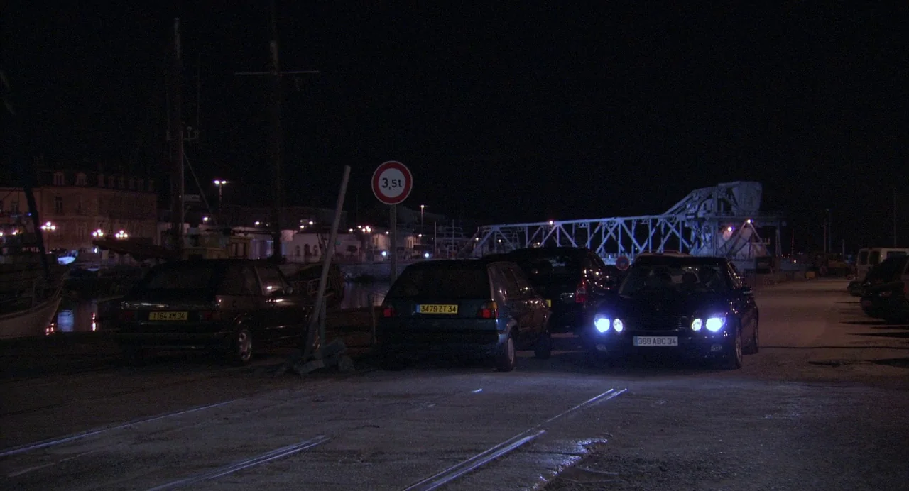 The Secret of the Grain (2007) movie still: wide — A dark harbor area features several parked cars next to a metal lift bridge. The scene is…