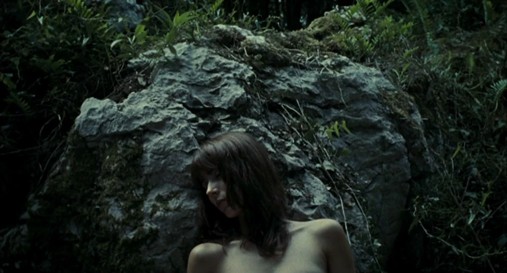 Timecrimes (2007) movie still: medium — A dense patch of woods features a prominent large grey boulder covered in moss. A young woman with long dark…