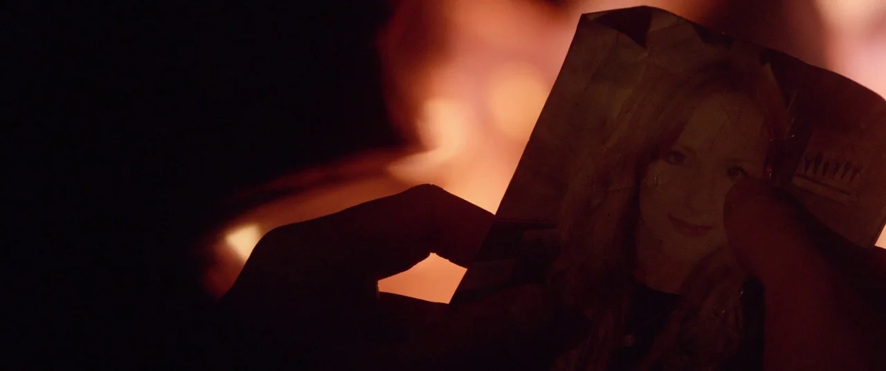 The Survivalist (2015) movie still: closeup — A hand holds a wrinkled photograph in front of a brightly burning campfire at night. The firelight…
