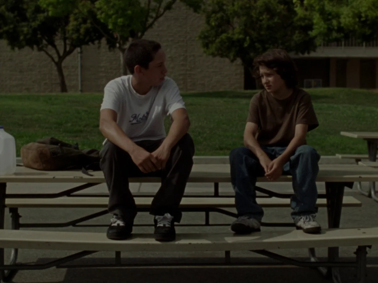 Mid90s (2018) movie still: medium — Two boys sit on a metal park table against a backdrop of trees. They sit together while facing one another on…