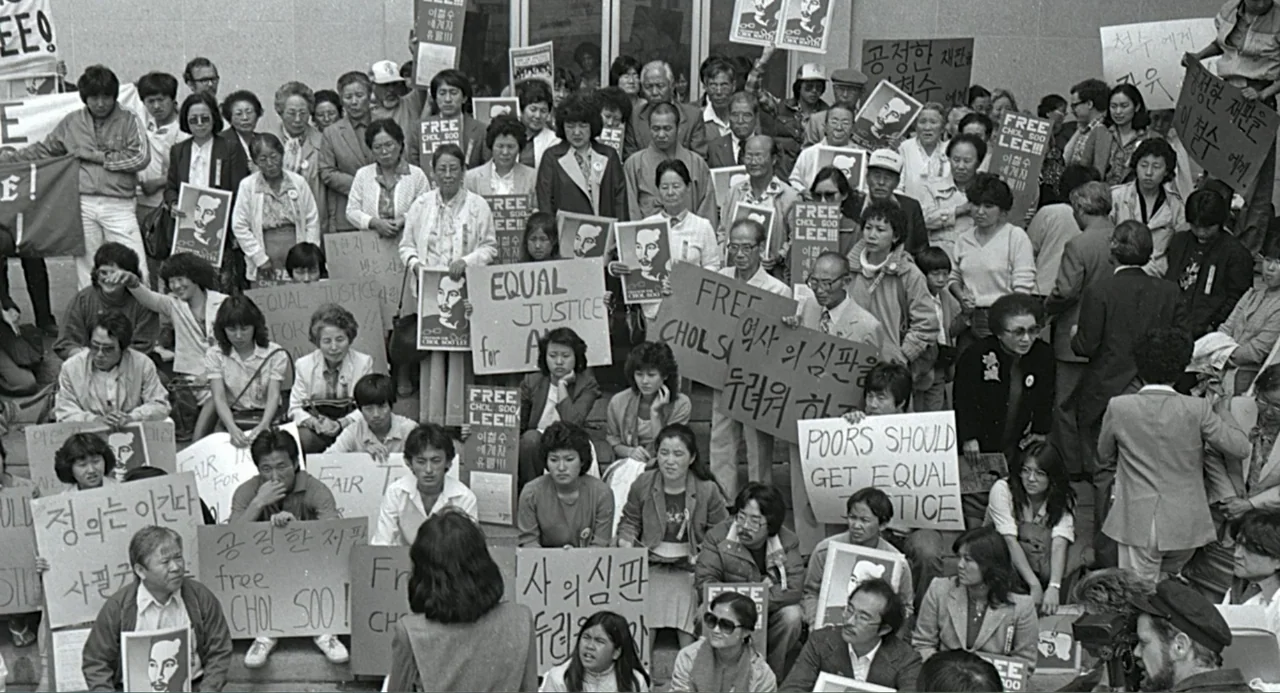 Free Chol Soo Lee (2022) movie still: wide — A large crowd of people gathers on wide stone steps outside a modern building. Participants hold…