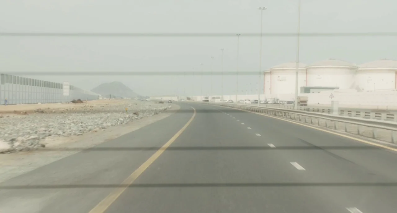 Bastards (2013) movie still: establishing — An asphalt highway curves through a dry, industrial landscape under a hazy sky. Large cylindrical…