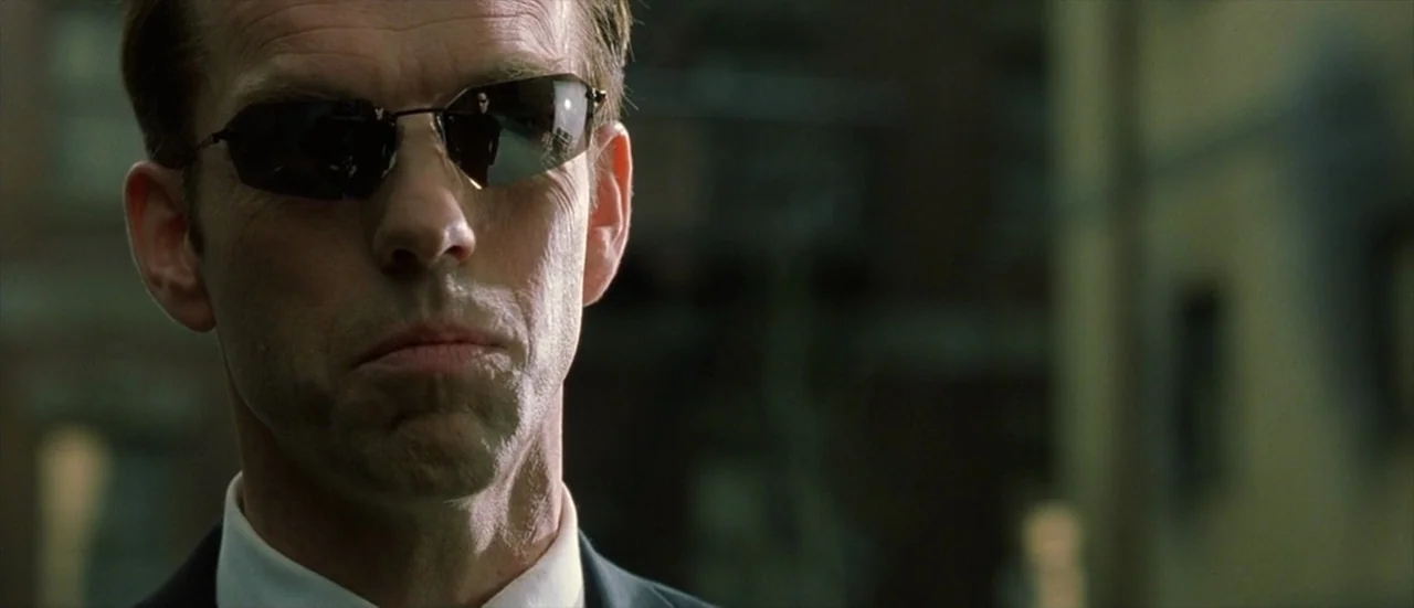 The Matrix Reloaded (2003) movie still: closeup — A man stands outdoors with an out-of-focus urban background visible behind him. The man maintains…