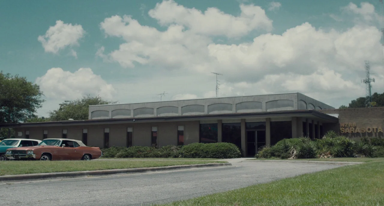 Christine (2016) movie still: establishing — A low-slung, mid-century commercial building with brick walls and a large parking lot. A vintage tan…
