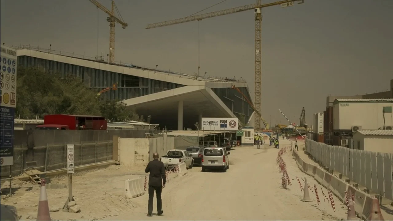 REM (2019) movie still: wide — A sprawling construction site features large yellow cranes and an unfinished modern building structure. A lone man in…